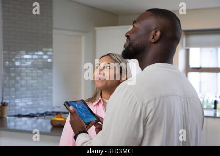 Afroamerikanischer Mann hält ein Tablet und eine hispanische Frau steht in der Küche zu Hause und trägt eine rosa Bluse Stockfoto