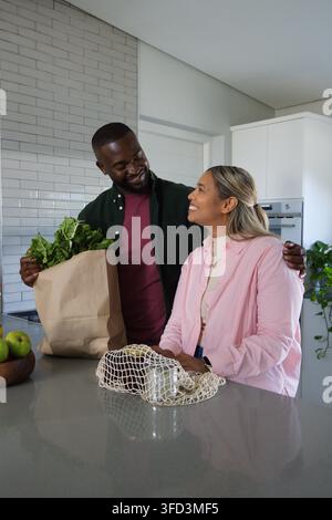 Afroamerikanisches Paar steht an einer glänzenden grauen Inselarbeitsplatte, Mann hält eine Papiereinkaufstasche Stockfoto