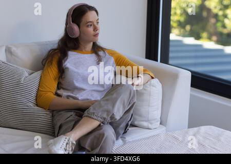 Frau Mitte 20, die auf einem hellen Ecksofa nahe einem großen Fenster sitzt, mit rosa Kopfhörern Stockfoto