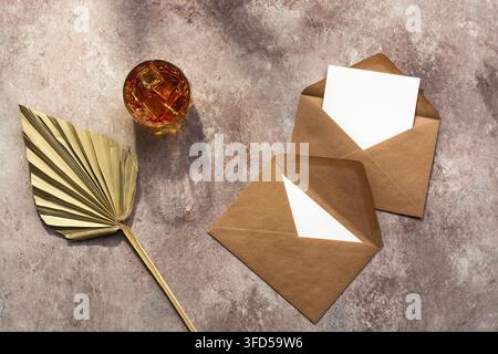 Whisky mit Eis in einem Glas, leere Papier-Mockup-Karten in einem Bastelumschlag, trockener Palmenblattfächer im Sonnenlicht. Männlicher Hintergrund. Vorlage für die Alkoholindustrie. Top-Down-Ansicht, Flat Lay. Beige grunge Hintergrund Stockfoto
