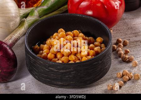 Gekochte Kichererbsen in einer schwarzen Keramikschale, umgeben von frischer roter Zwiebel, Knoblauch, grünen Zwiebeln, roter Paprika auf einer strukturierten Oberfläche Stockfoto