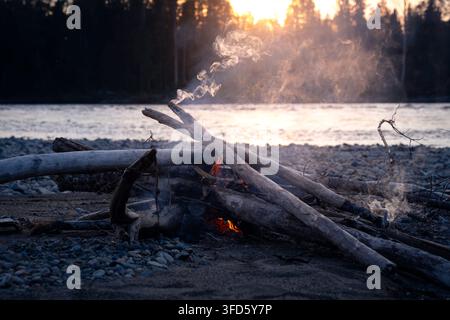 Lagerfeuer brennt zwischen Treibholzblöcken an einem felsigen Flussufer mit ruhigem Wasser und Wald bei Sonnenuntergang Stockfoto
