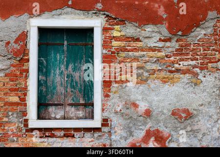 Rustikale grüne Holzläden sitzen in einem weißen Steinrahmen an einer Wand, wo verblasster Terrazzoputz abgeblättert ist, um raues rotes Backstein darunter freizulegen. Stockfoto