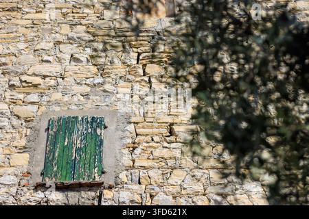 Alte Steinmauer eines rustikalen Gebäudes mit einem kleinen Fenster links, dessen verwittertes grünes Holzfensterläden sich ablösen und sonnenverblichen sind. Stockfoto