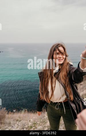 Frau Klippen Ozean lächelnde Frau mit langen Haaren steht auf einer Klippe mit Blick auf ein türkisfarbenes Meer an einem windigen Tag Stockfoto
