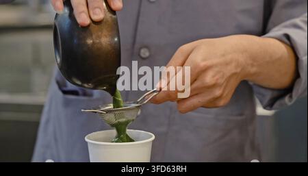 Barista-Hände gießen grüne Matcha-Flüssigkeit durch Metallsiebe in einen weißen Pappbecher, professionelle Getränkezubereitung in der Café-Küche, kulinarisches Dokument Stockfoto