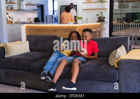 Afroamerikanische Familie unterhält sich in der Nähe der Kücheninsel, während zwei Jugendliche auf dem Sofa ein Tablet teilen Stockfoto