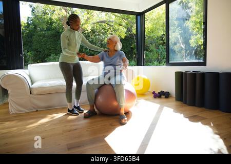 Seniorin und afroamerikanische Frau üben das Gleichgewicht, sitzen auf einem rosa Stabilitätsball zu Hause Stockfoto