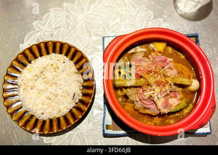 Japanisches Suppencurry serviert in einem roten Tongefäß mit geschnittenem Fleisch, Gemüse und einem Beilagenteller mit Mischungsreis. Stockfoto