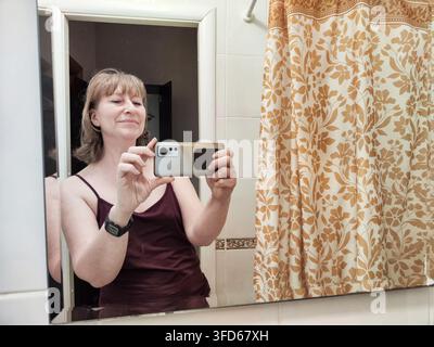 Eine Frau steht vor einem Spiegel in ihrem Badezimmer und macht ein Selfie mit einem Smartphone. Sie lächelt und fühlt sich in ihrer Umgebung wohl. Stockfoto