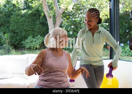 Seniorin und Trainer helfen einer sitzenden Frau in einem lilafarbenen Oberteil mit violetten Hanteln im Wohnzimmer Stockfoto