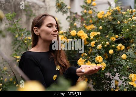 Junge Frau genießt fallende Rosenblätter im Freien, umgeben von schönen gelben Blüten im Frühling Stockfoto