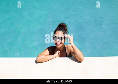 Lächelnde Frau trägt eine Sonnenbrille und entspannt sich in einem Schwimmbecken an einem sonnigen Sommertag, mit türkisfarbenem Wasser und großzügigem Kopfraum. Helles Lifestyle-Foto Stockfoto