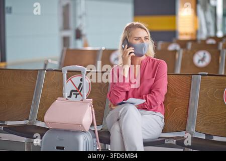 Eine Frau mit einer Gesichtsbedeckung spricht am Handy, während sie in einem Wartebereich des Flughafens sitzt, mit Schildern, die Abstandsregeln anzeigen Stockfoto