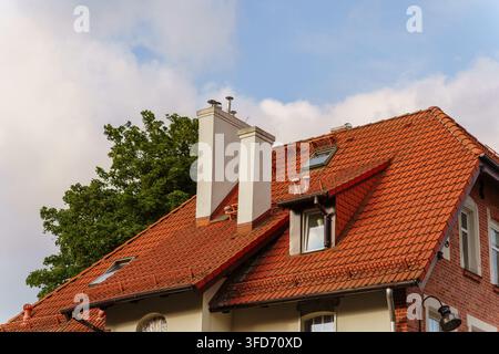 Rotziegel-Dach auf einem traditionellen Wohnhaus mit hohen weißen Schornsteinen und Dachfenster unter einem blauen bewölkten Himmel, architektonisches Außendetail Stockfoto