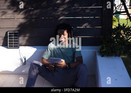 Mann sitzt auf eingebauter Betonbank in beschatteter Terrasse und hält Smartphone in der Nähe einer Solarplatte Stockfoto
