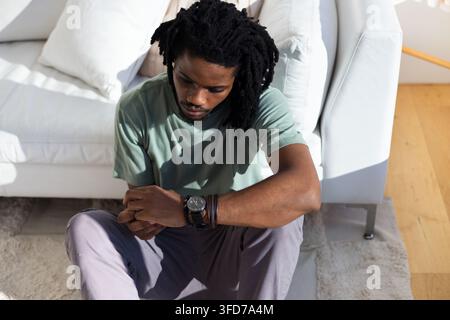 Erwachsener afroamerikanischer Mann sitzt auf einem Teppich im Wohnzimmer, lehnt sich an ein Sofa und zeigt Uhr und Armbänder Stockfoto