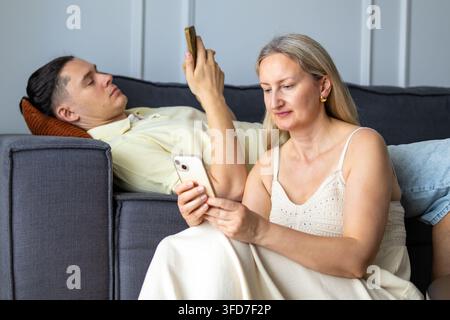 Mittlere Frau sitzt neben Mann, während beide auf ihrer Couch zusammen auf ihre eigenen Telefone schauen. Konzept der gemeinsamen Bildschirmzeit und parallelen Telefonnutzung während des Putsches Stockfoto
