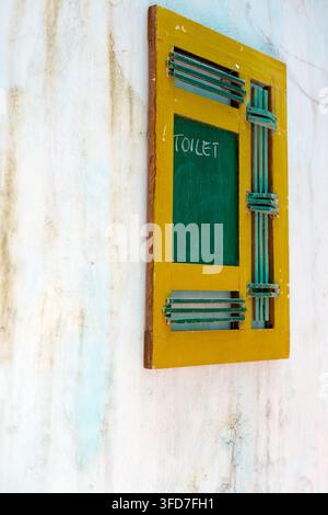 Ein kreativer gelber Holzrahmen für Fensterläden, montiert an einer verwitterten, strukturierten Wand mit dem Wort "TOILET" in Kreide geschrieben Stockfoto