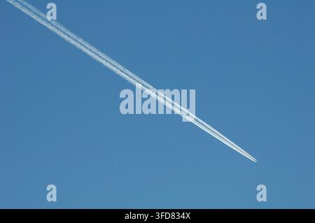 Hochflugzeug, das diagonal über einen klaren blauen Himmel fliegt und zwei deutliche weiße Kondensstreifen hinterlässt, die atmosphärische Bedingungen verdeutlichen Stockfoto