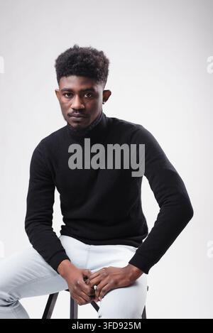 Ein stilvoller junger Mann mit dunkler Haut und einer Afro-Frisur posiert selbstbewusst im Sitz gegen einen schlichten Hintergrund Stockfoto