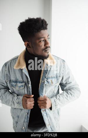 Ein stilvoller junger schwarzer Mann mit Afro-Frisur posiert selbstbewusst in einer hellblauen Jeansjacke über einem schwarzen Rollkragenpullover Stockfoto