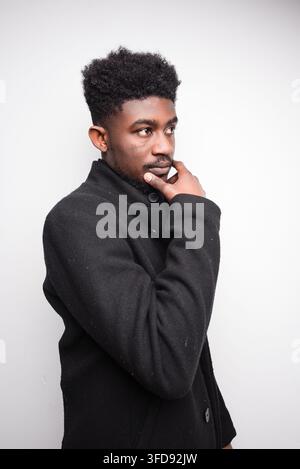 Ein junger Mann mit Afro-Frisur und Bart denkt nach, während er einen dunklen Mantel trägt, vor einem schlichten Hintergrund Stockfoto