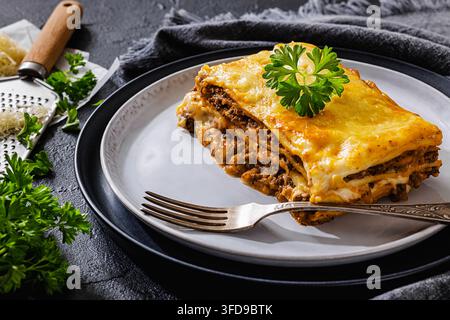 Portion Fleischlasagne mit Hackfleisch, Tomatensoße und goldener Cheddar-Kruste, serviert auf einem grauen Keramikteller mit einer Gabel auf einer schwarzen Tischplatte, keine Stockfoto
