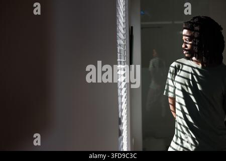 Afroamerikaner in grünem T-Shirt, steht am Fenster mit Jalousien, Spiegelreflexion, Kopierraum Stockfoto