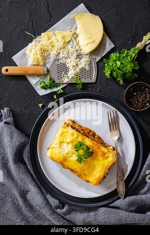 Gebackenes Lasagnequadrat mit cremiger Sauce, geschmolzener Käseschicht und Rinderragout, serviert auf einem grauen Teller neben einer Reibe auf einem schwarzen Tisch, Nahaufnahme, verti Stockfoto