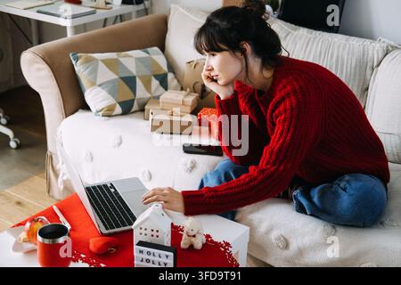 Frau in gestricktem Pullover arbeitet an einem Laptop auf dem Sofa neben verpackten Paketen und einem HOLLY JOLLY-Schild. Remote-Arbeit Dezember, Laptop-Heimbüro Weihnachten Stockfoto