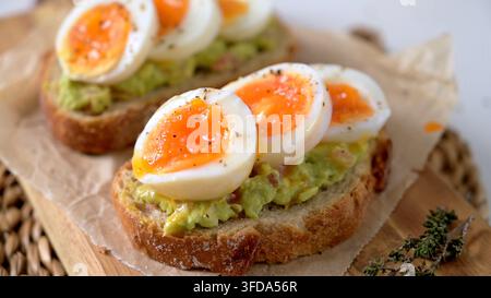 Zwei Scheiben knuspriges Sauerteigbrot mit Avocadopüree und perfekt weich gekochten Eiern mit laufendem Eigelb. Die natürliche Seitenbeleuchtung unterstreicht den Stockfoto