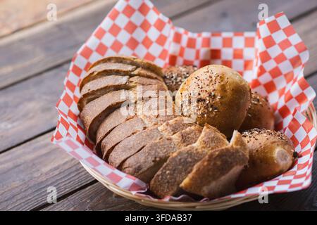 Korb gefüllt mit geschnittenem handwerklichem Sauerteigbrot und frisch gebackenen gesäten Brötchen auf einem rustikalen Holztisch. Hausgemachtes Weizensauerteigbrot, wunderschön Stockfoto