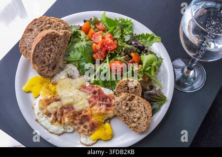 Ein köstliches und ausgewogenes Frühstück, das auf einem weißen Teller auf einem dunklen Tisch serviert wird. Das Gericht enthält gebratene Eier mit geschmolzenem Käse und knusprigem Speck, ein Stockfoto