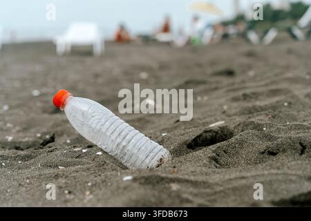 Verlassene Plastikflasche auf einem dunklen Sandstrand. Benutzte Wasserflasche aus Plastik mit rotem Verschluss, halb vergraben im dunklen Vulkansand eines Strandes, mit verschwommenem Hintergrund aus Ozean und entfernten Strandbesuchern. Stockfoto