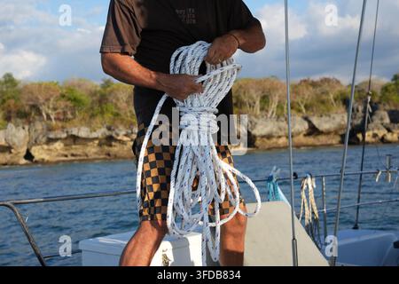 Ein Seemann wickelt ein weißes Nylonseil in seinen Händen auf dem Deck eines Segelboots vor der Kulisse einer felsigen Küste und des blauen Meeres Stockfoto