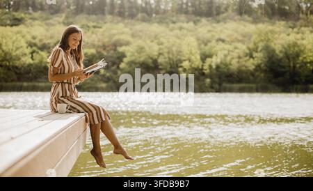 Eine junge Frau genießt ein Buch am Steg, ihre Füße tauchen in das ruhige Wasser unter einer hellen Sonne. Stockfoto