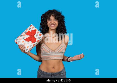 Fröhliche junge Frau hält eine Geschenkschachtel, lächelt breit auf blauem Hintergrund Stockfoto