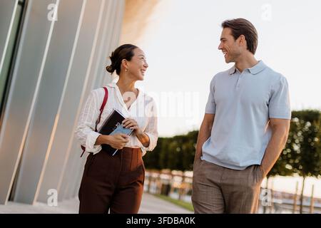 Eine koreanische Frau und ein kaukasischer Mann lächeln sich an, während sie einen hellen Abend zusammen in der Nähe moderner Architektur am Wasser genießen. Stockfoto