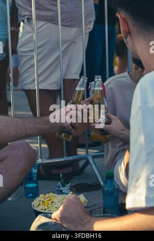 Nahaufnahme von Freunden, die Corona-Bierflaschen neben Metallabsperrungen für Menschenmassen auf einem öffentlichen Freiluftfest anstoßen, mit Popcorn im Vordergrund Stockfoto