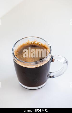 Frisch gebrühter schwarzer Espresso mit cremigem Schaum, serviert in einer transparenten Glastasse, isoliert auf einer hellen weißen minimalistischen Tischfläche. Stockfoto