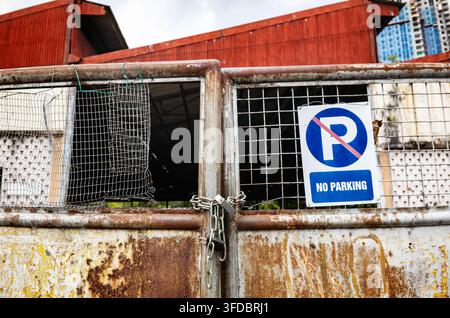 Ein industrielles Straßenfoto mit einem gealterten, rostigen Metalltor, das mit einer schweren Kette und einem Vorhängeschloss gesichert ist und ein auffälliges "No Parking"-Schild zeigt Stockfoto