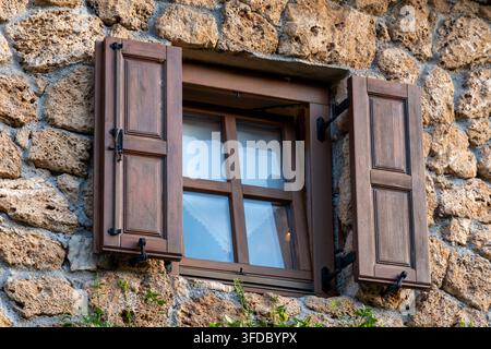 Ein hölzernes Fenster mit offenen Läden ist in eine raue Steinmauer an der Fassade eines traditionellen Gebäudes eingelassen. Stockfoto