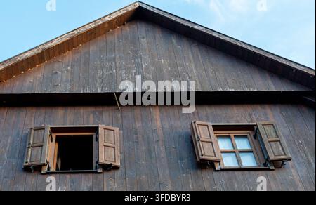 Eine verwitterte Holzhaustür mit zwei offenen Fenstern und Fensterläden steht vor einem klaren blauen Himmel. Stockfoto