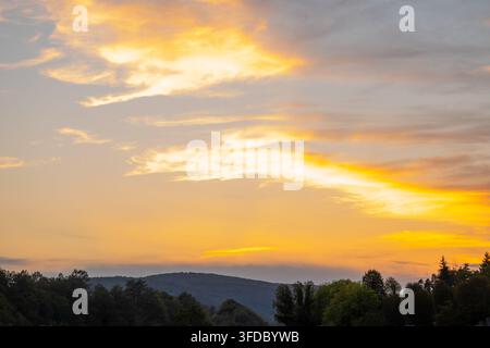 Goldene Sonnenuntergangswolken über bewaldeten Hügeln und einer ruhigen Abendlandschaft Stockfoto