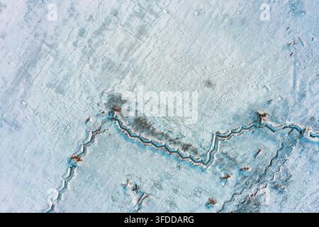 Spuren schweren Winterfrosts auf gefrorenem Boden und eisiger Oberfläche mit türkisfarbenen Mineralrissen und Schneeschicht, Luftansicht von oben Stockfoto