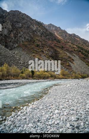Der türkise Alamedin-Fluss fließt über Felsen und Kieselsteine, flankiert von einem breiten, grauen felsigen Flussbett auf der rechten Seite und einem schmalen Streifen aus Gelb und Orange Stockfoto