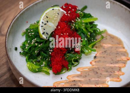 Eine Schüssel grüner Algensalat ist mit einem Streifen rotem Tobiko, Sesamsamen und einer Limettenscheibe garniert und mit einer Beilage aus Soße serviert. Stockfoto