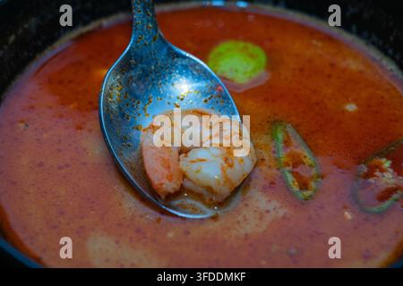 Ein Löffel hebt eine Garnele aus einer Schüssel mit reichhaltiger, würziger thailändischer Tom-Yum-Suppe. Stockfoto