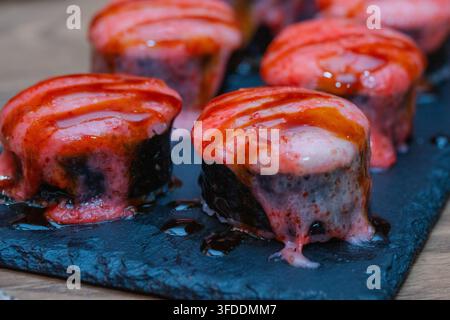 Eine Nahaufnahme zeigt mehrere gebackene oder angegrillte Sushi-Rollen, die mit einer rosa cremigen Sauce und süßer Sozuglasur belegt sind und auf einem dunklen Schieferteller arrangiert wurden. Stockfoto
